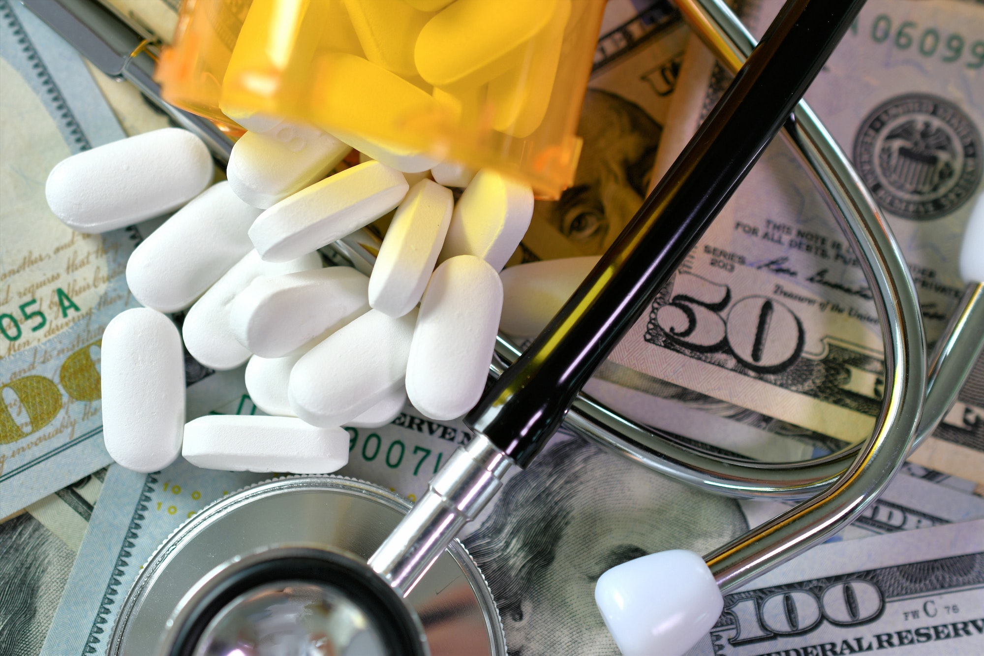 A stethoscope laying on medicine white pills and money with an RX prescription drug bottles