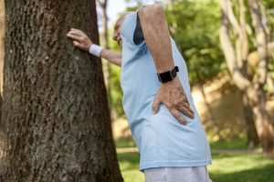 Senior man suffers from pain in lower back leaning onto tree at training in park