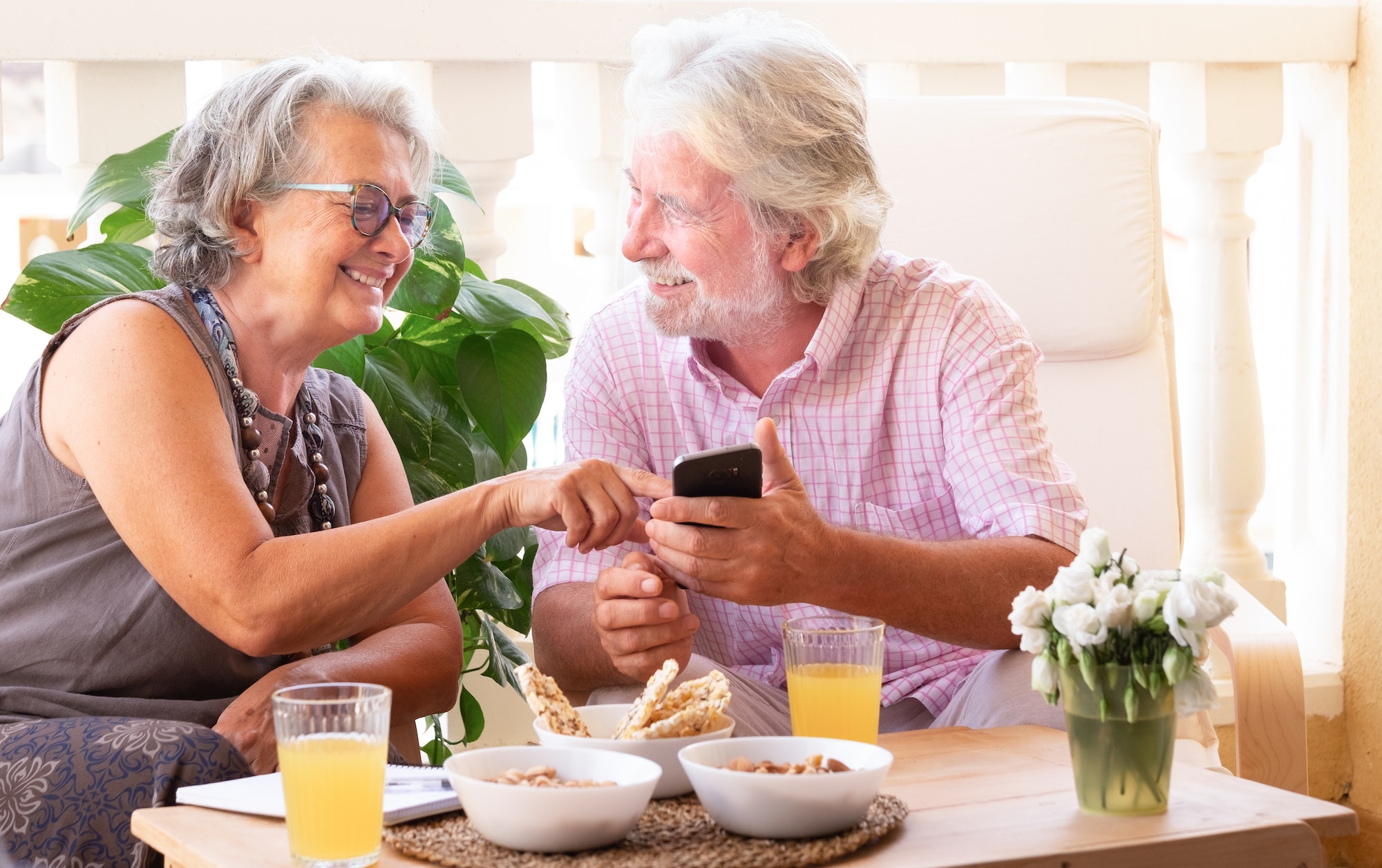 Two senior people pensioners with white and gray hair looking at the same cell phone enjoying snacks